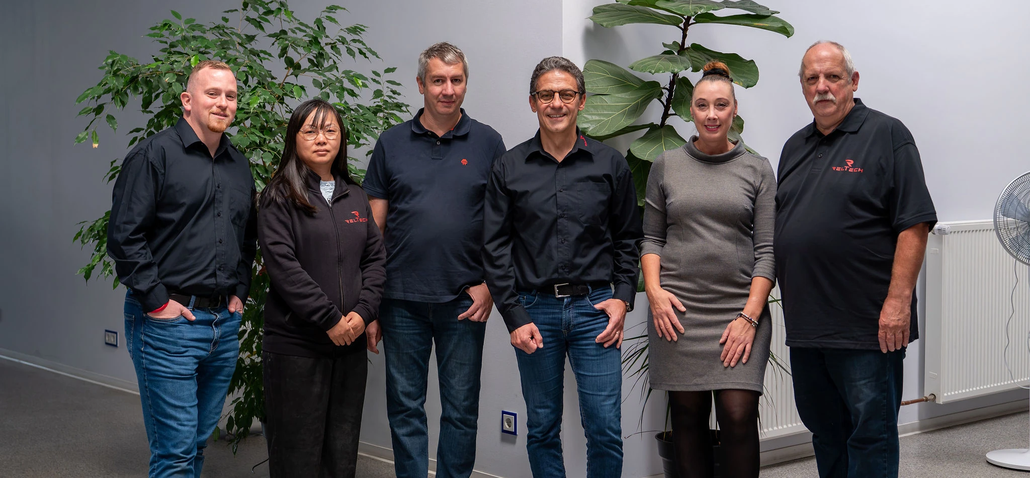 Gruppenfoto von sechs Mitarbeitenden des Unternehmens RelTech, die nebeneinander in einem hellen Büro mit großen Grünpflanzen stehen.